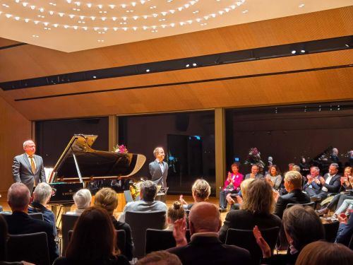 Two gentlemen in suits stand at a black grand piano and look out into the clapping audience