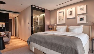 Einblick in das Deluxe Zimmer mit großem Doppelbett unter abstrakten Gemälden mit direktem Zugang zum Badezimmer mit Glastür