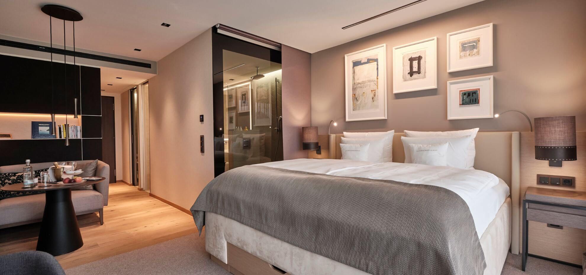 Einblick in das Deluxe Zimmer mit großem Doppelbett unter abstrakten Gemälden mit direktem Zugang zum Badezimmer mit Glastür